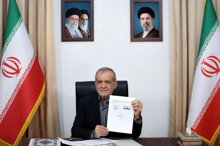 الرئيس الإيراني مسعود بزشكيان يجلس خلف مكتب، ممسكًا بمذكرة تفاهم، مع العلم الإيراني خلفه وصور لقيادات إيرانية.