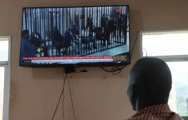 شخص يشاهد بثًا مباشرًا لمحاكمة رياك مشار، النائب الأول لرئيس جنوب السودان، على شاشة تلفاز في قاعة محكمة، مع وجود حواجز أمنية.