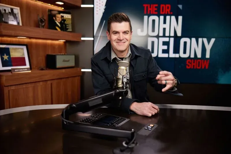 الدكتور جون ديلوني يتحدث إلى الكاميرا في استوديو بودكاست، مرتديًا قميصًا أسود، مع ميكروفون وأجواء مريحة تعكس موضوعات الصحة النفسية.