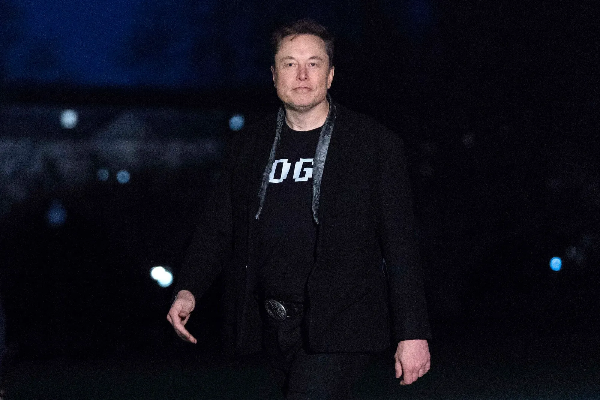 إيلون ماسك يسير في الليل مرتديًا قميصًا أسود مكتوب عليه "DOGE"، مع خلفية مظلمة تعكس أجواء العمل والابتكار.