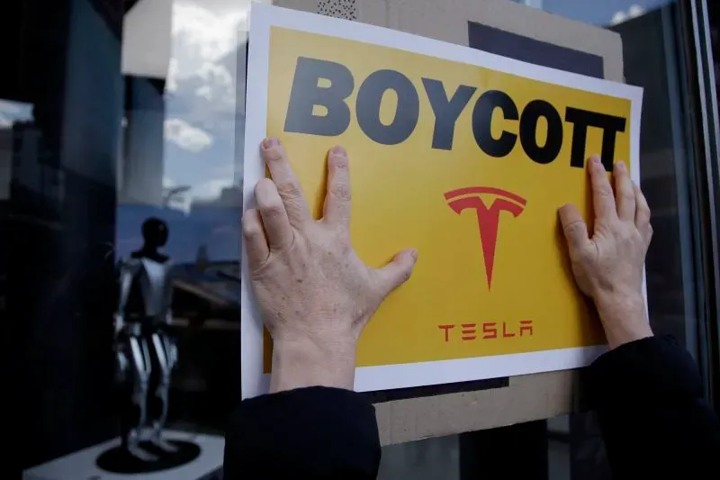 لافتة كبيرة مكتوب عليها "مقاطعة تسلا" بلون أصفر، تُثبتها يد، مع وجود تمثال إنسان آلي في الخلفية، تعكس دعوات لمقاطعة الشركة.