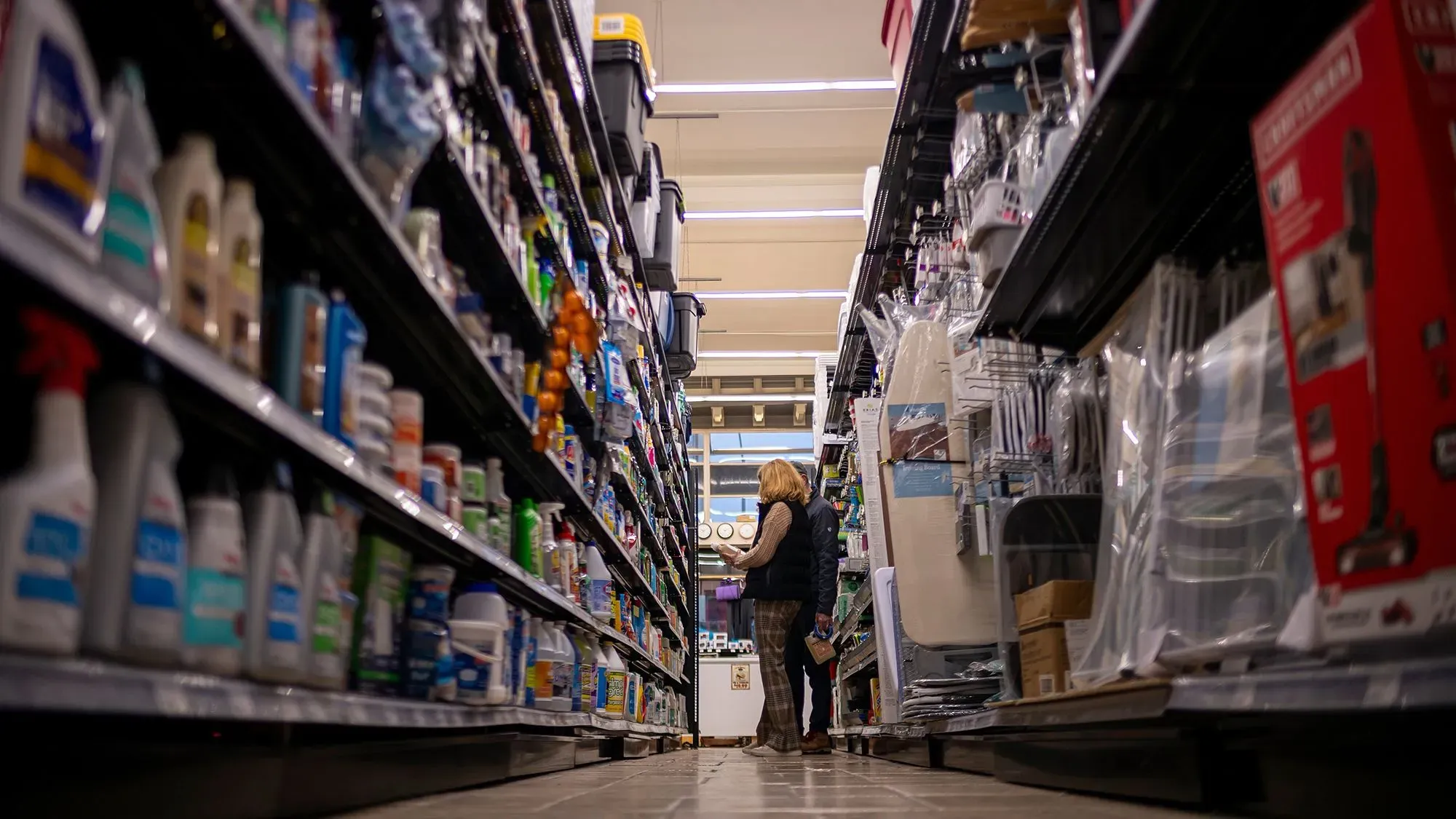 امرأة تتسوق في ممر متجر، محاطة بمنتجات تنظيف وأدوات منزلية، تعكس ارتفاع الأسعار والتضخم في السوق الأمريكية.