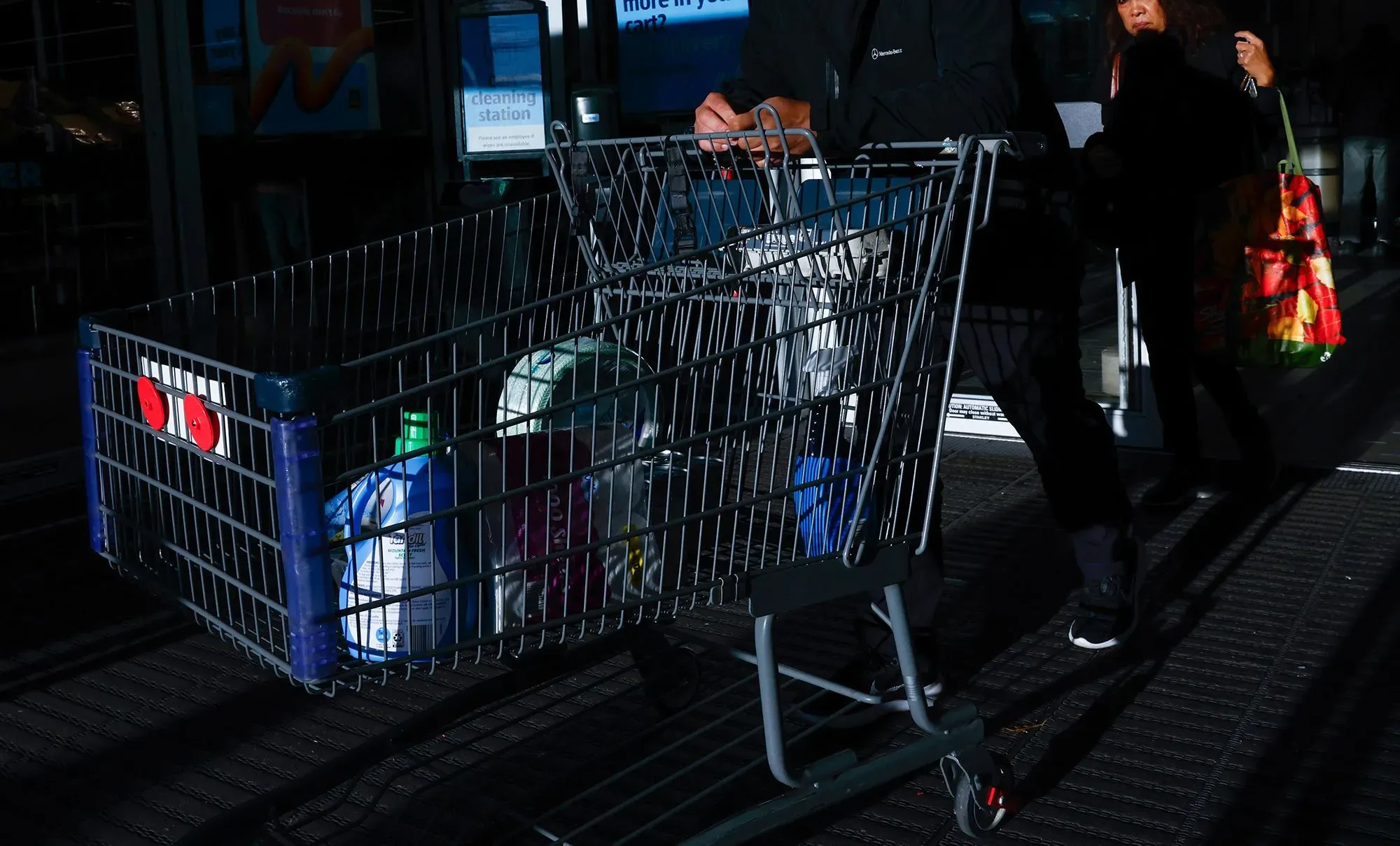 سلة تسوق تحتوي على منتجات متنوعة، تُظهر تأثير التضخم على الأسعار، بينما يمر شخصان بالقرب من المدخل.