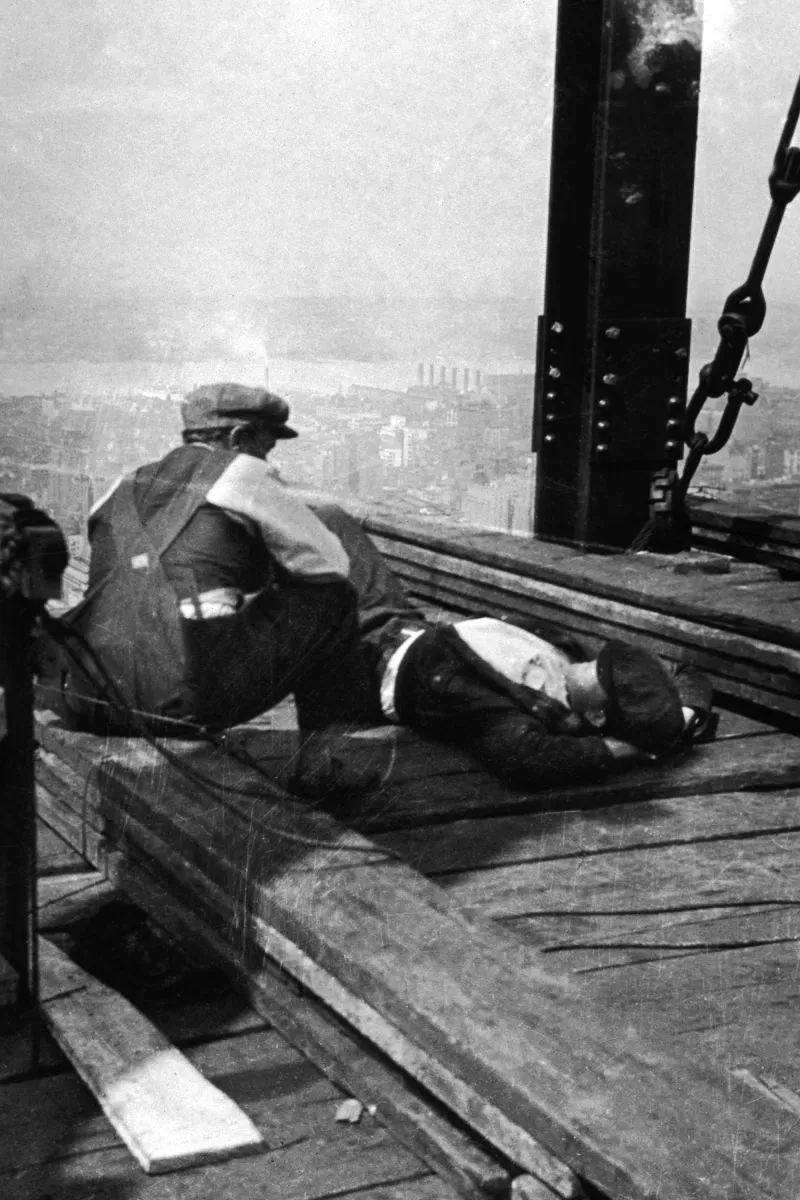 عمال بناء يجلسون على هيكل مبنى كرايسلر في نيويورك، مع إطلالة على المدينة في الخلفية، خلال فترة بناء المبنى في الثلاثينيات.