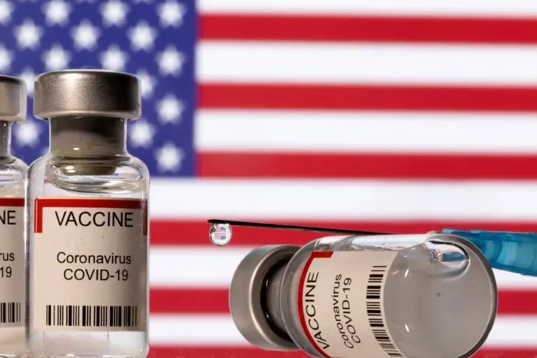 زجاجات لقاح كوفيد-19 مع حقنة أمام علم الولايات المتحدة، تعكس الجدل حول مكونات اللقاحات مثل الألمنيوم وتأثيراتها.