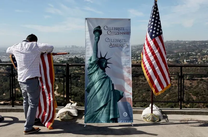 شخص يحمل علم الولايات المتحدة أمام لافتة تحتفي بالجنسيات، مع وجود علم آخر في الخلفية، يُظهر رمز الحرية.