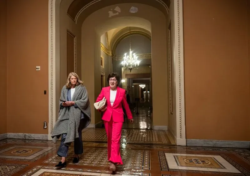 امرأتان تسيران في ممر مزين بأضواء كريستالية، واحدة ترتدي بدلة وردية والأخرى تحمل بطانية، مما يعكس أجواء الكونغرس.