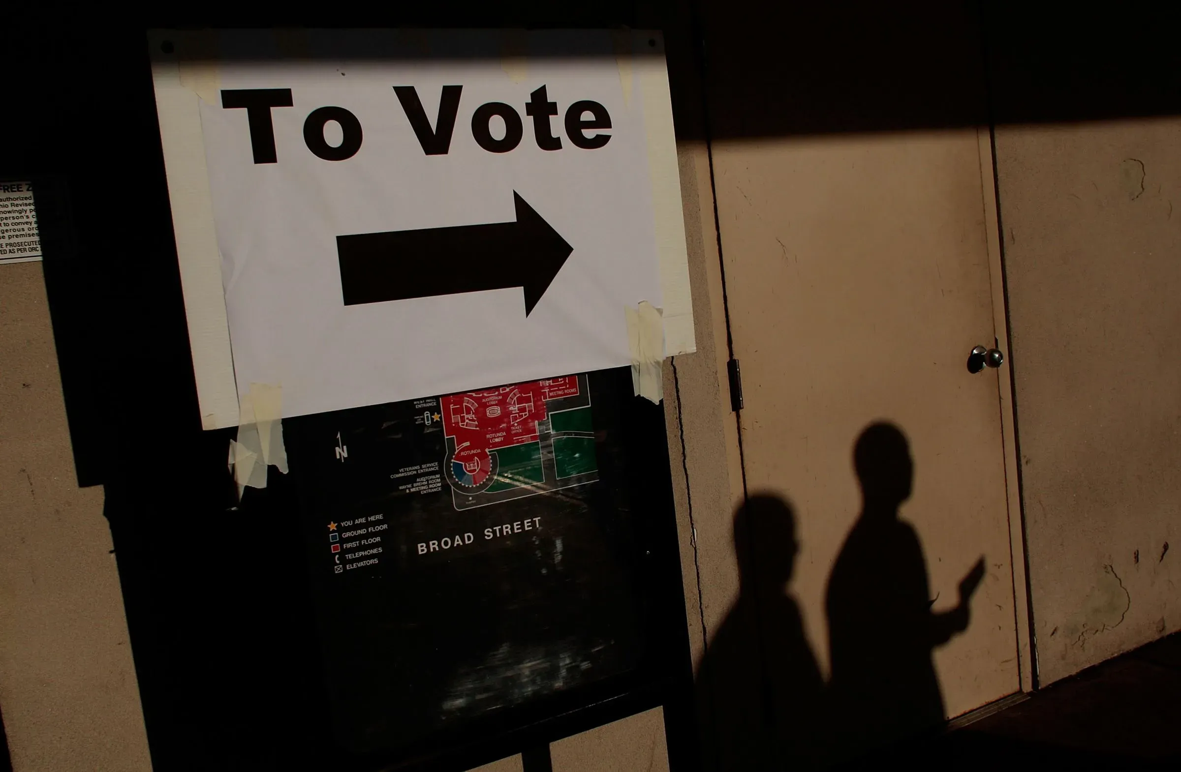 علامة تشير إلى اتجاه الاقتراع مع سهم يشير إلى اليمين، وظلال شخصين في الخلفية، تعكس أهمية المشاركة في الانتخابات.