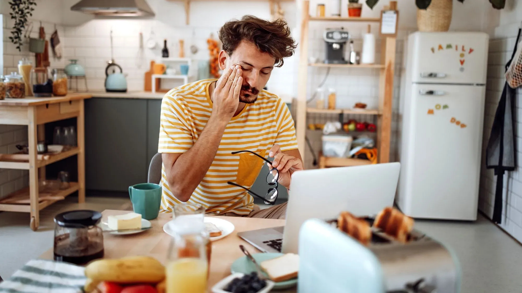 رجل يشعر بالتعب والإرهاق أثناء تناول الإفطار في المطبخ، محاطًا بأدوات المطبخ ومكونات الطعام، بينما يعمل على الكمبيوتر المحمول.