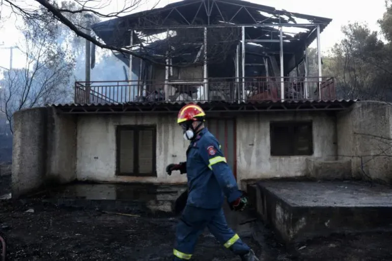 رجل إطفاء يرتدي معدات الحماية يسير بجانب منزل محترق جزئيًا في منطقة تأثرت بحرائق الغابات في اليونان.