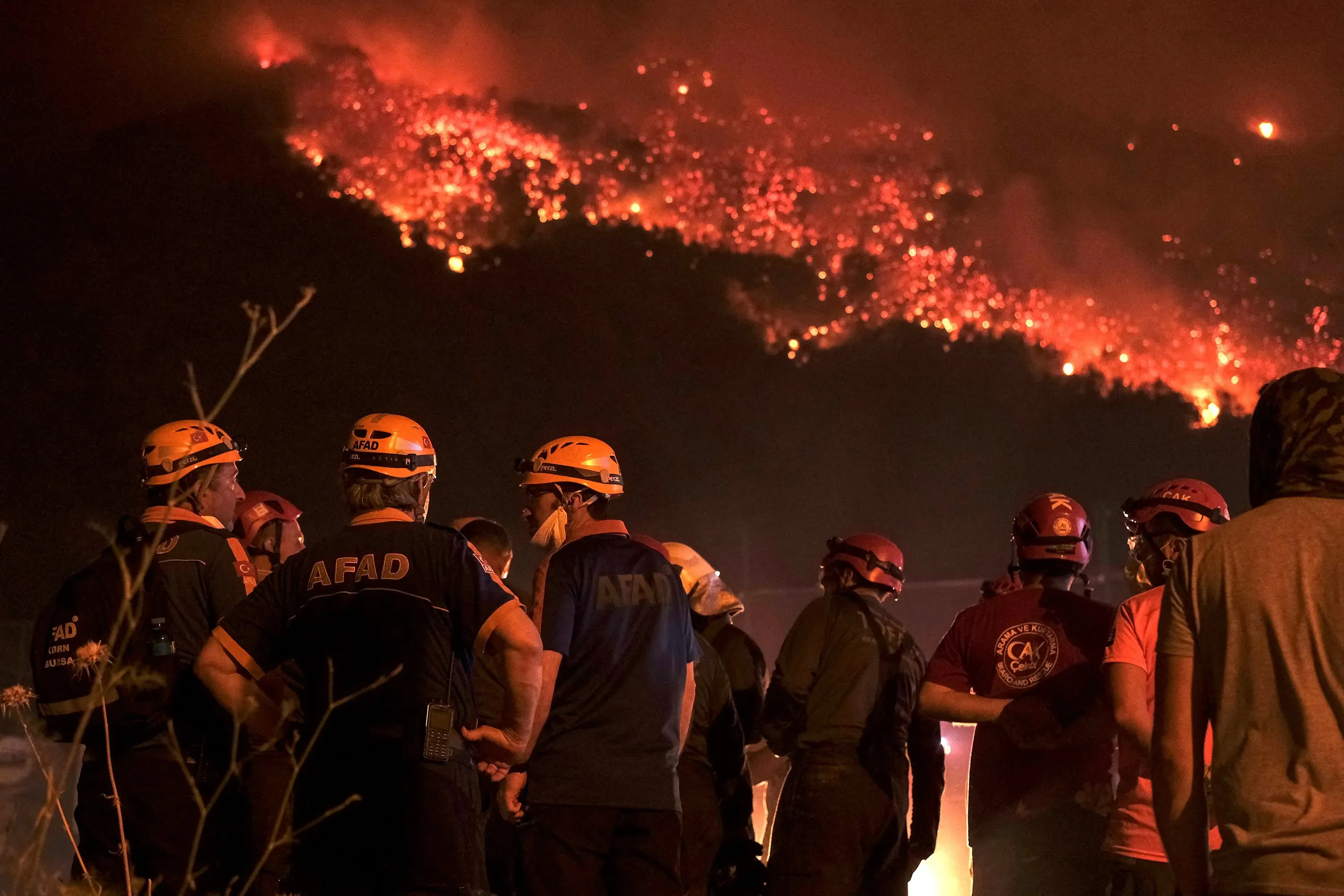 رجال إطفاء يرتدون خوذات، يتجمعون لمواجهة حرائق الغابات المشتعلة في الخلفية، حيث تتوهج السماء باللون الأحمر بسبب النيران.
