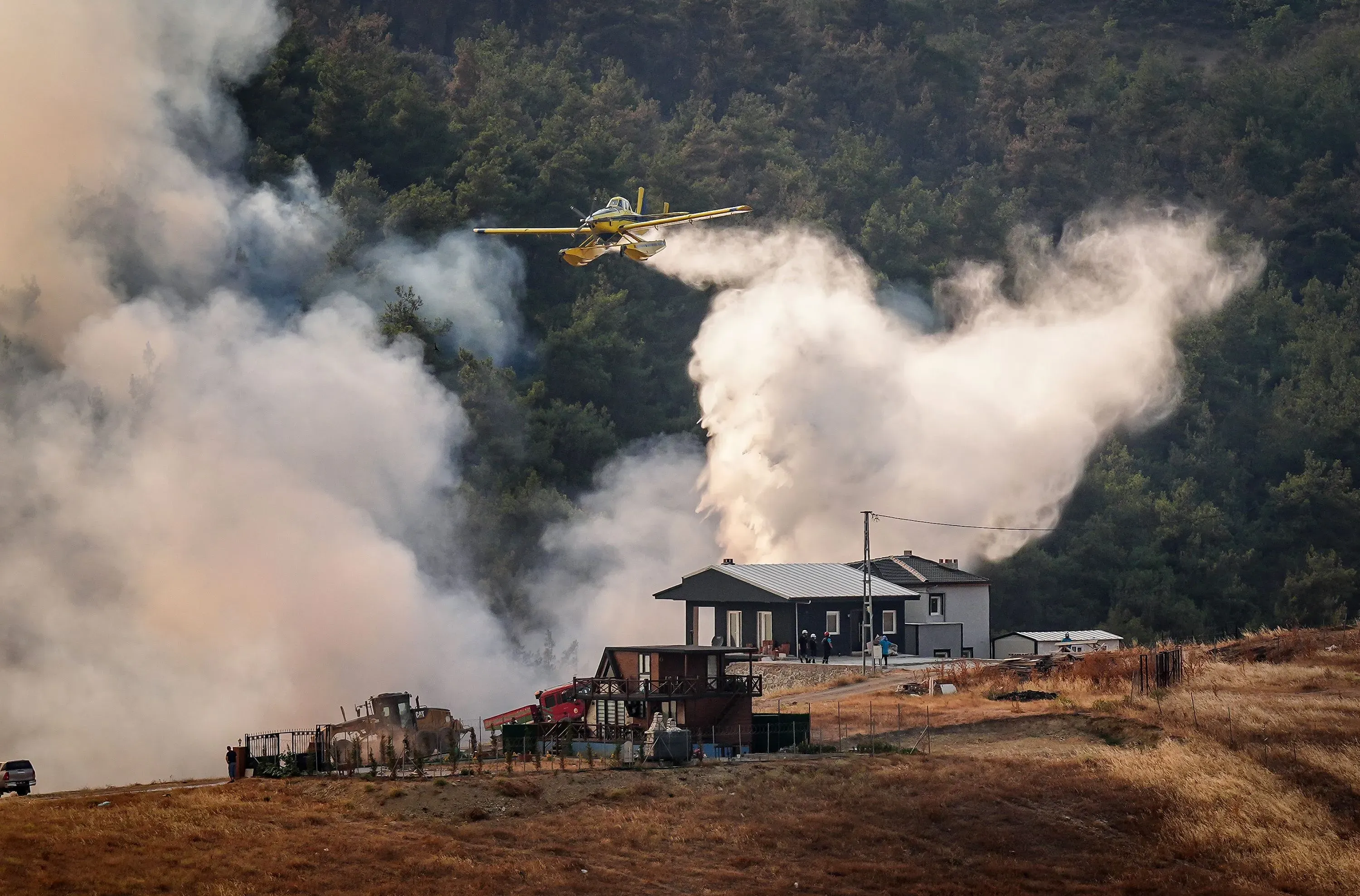 طائرة إطفاء تحلق فوق منطقة مشتعلة، تطلق المياه على النيران التي تهدد المنازل في تركيا، وسط سحب من الدخان الكثيف.