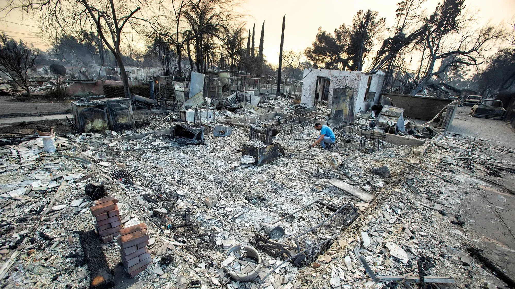 رجل يجمع الأنقاض من منزل دمرته حرائق الغابات في لوس أنجلوس، مما يسلط الضوء على تأثير الكوارث الطبيعية على المجتمعات.