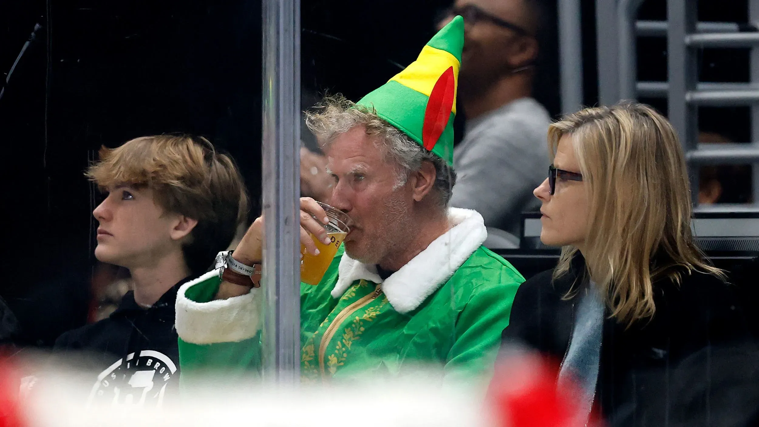 ويل فيريل يرتدي زي بادي العفريت في مباراة هوكي، يحتسي مشروبًا مع تعبير جاد، محاطًا بأشخاص آخرين.