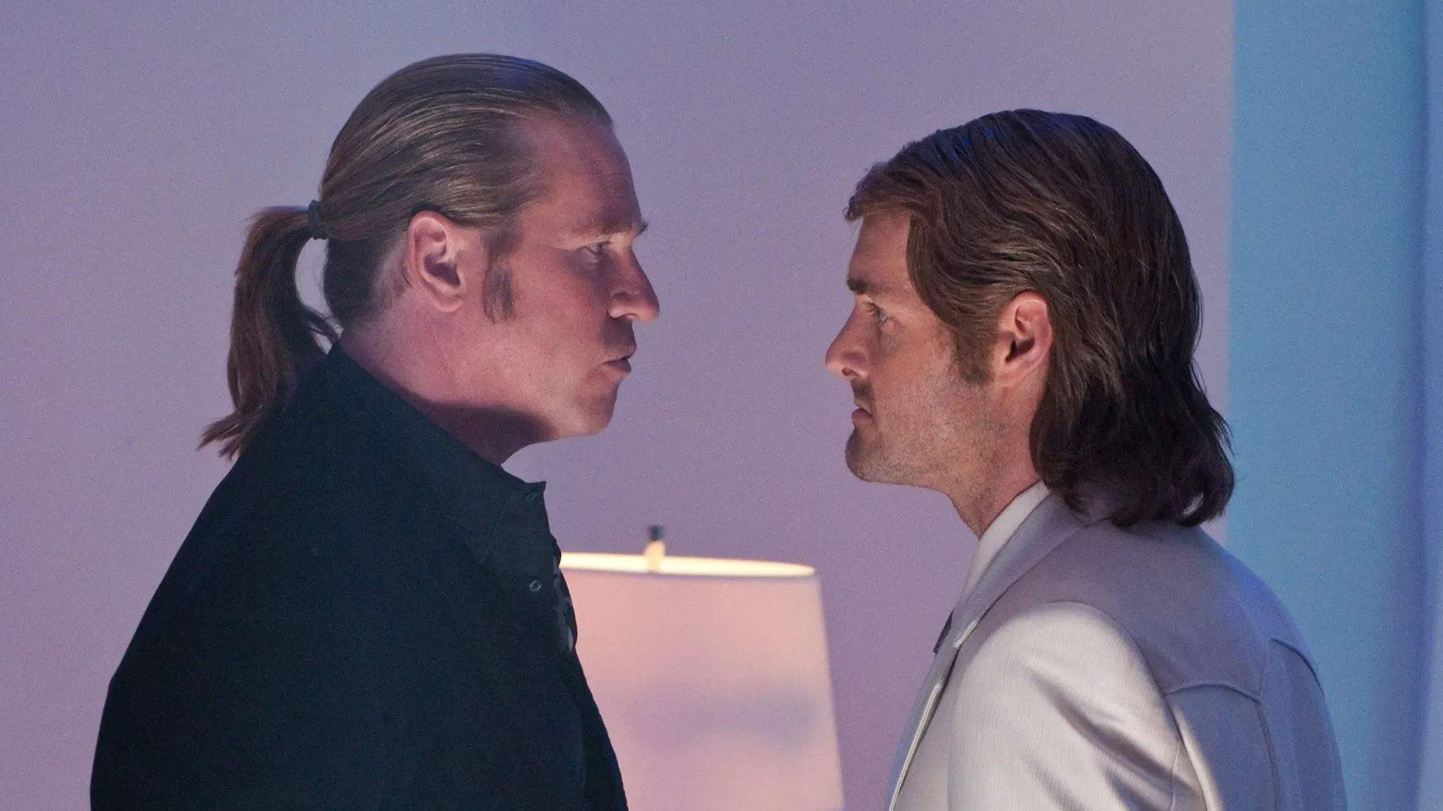 تظهر الصورة مواجهة بين شخصين، أحدهما يرتدي ملابس داكنة والآخر يرتدي بدلة بيضاء، مما يعكس التوتر بين شخصيتين في مشهد درامي.