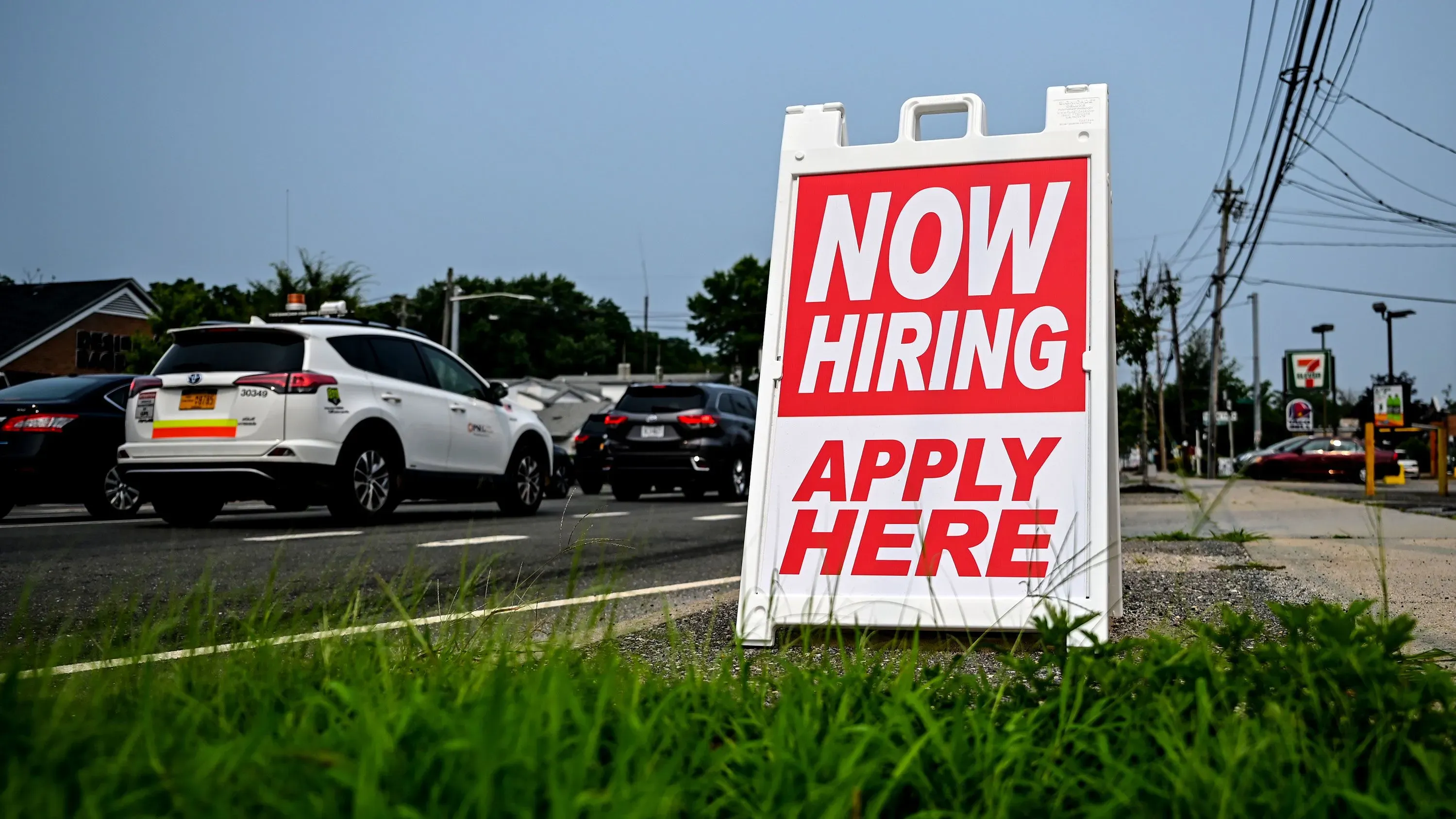 لافتة كبيرة مكتوب عليها "نحن نوظف الآن" مع تفاصيل التقديم، تظهر على جانب الطريق وسط حركة مرور نشطة.