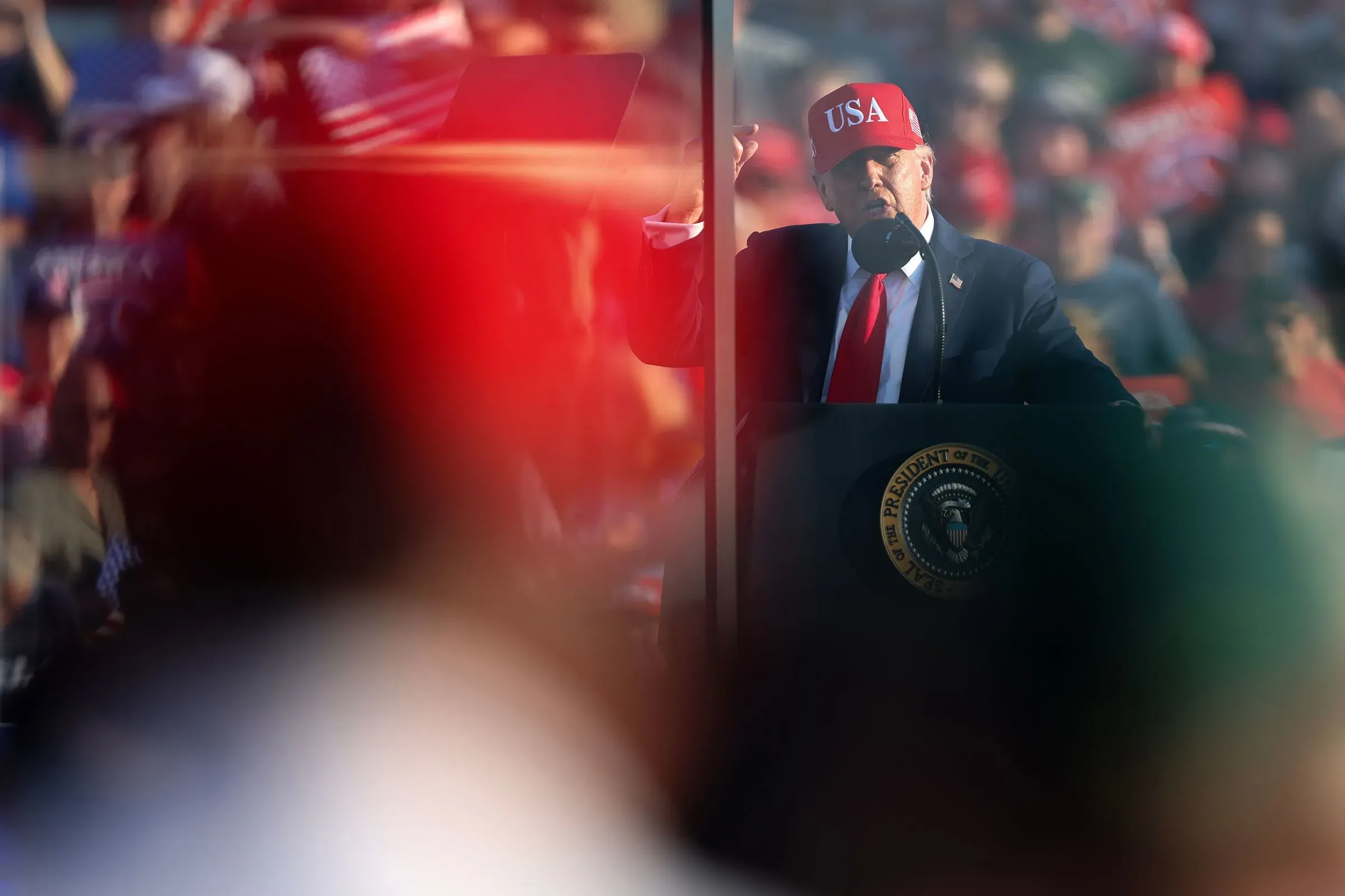 دونالد ترامب يتحدث خلال تجمع انتخابي، مرتديًا قبعة تحمل كلمة "USA"، مع وجود حشد خلفه، مما يعكس أجواء الحملة الانتخابية.