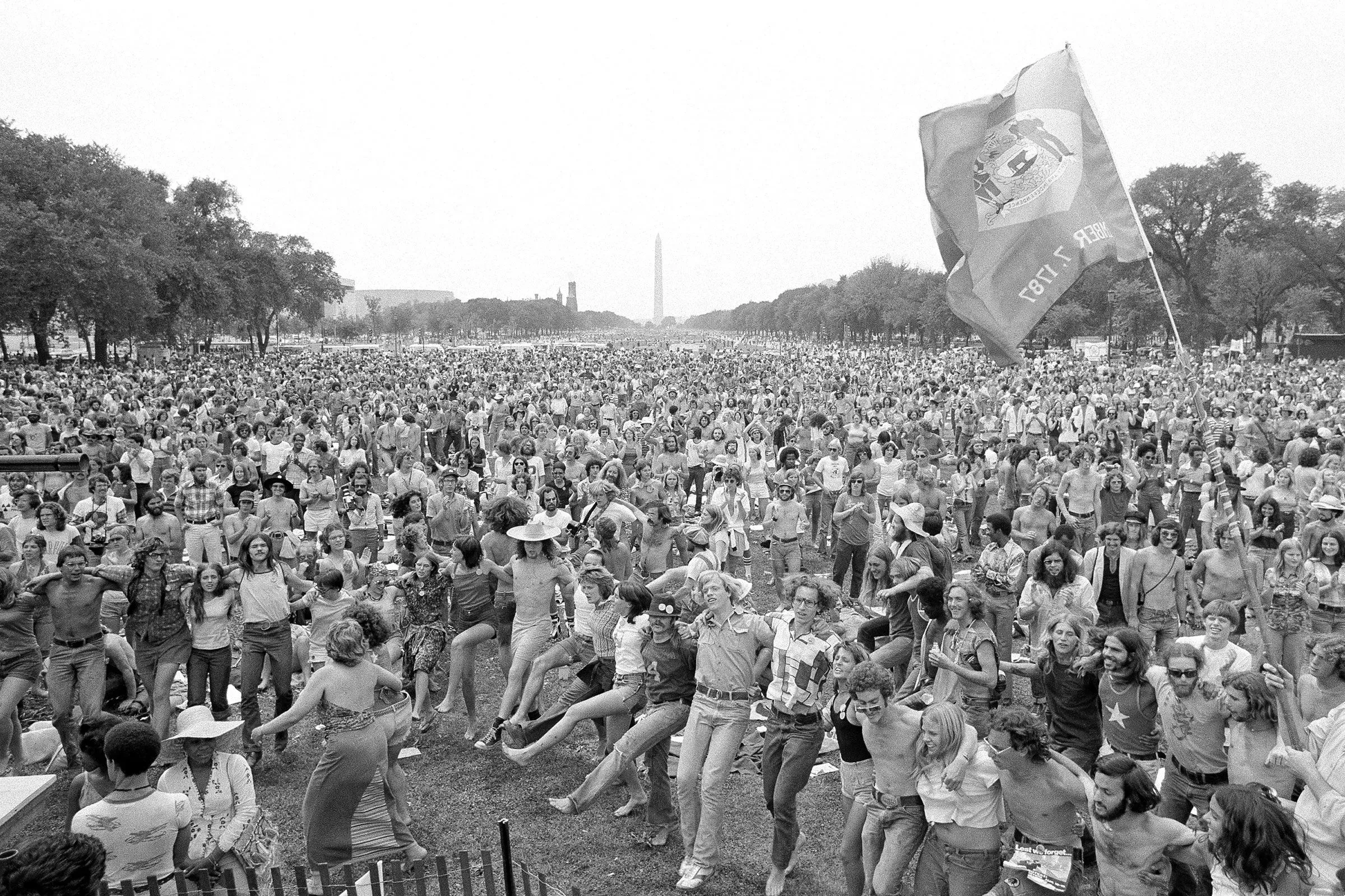 حشود كبيرة تحتفل في تجمع جماهيري، مع علم يرفرف في المقدمة، ومشهد لتمثال واشنطن في الخلفية، يعكس روح الوطنية.