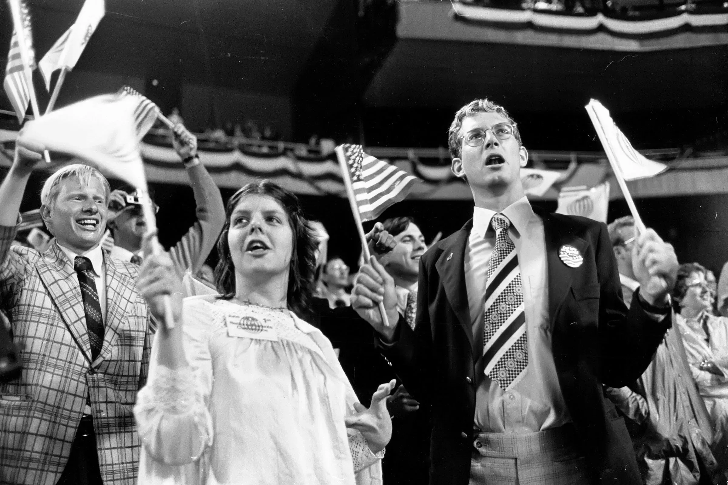 مجموعة من الأشخاص في حدث جماهيري يلوحون بالأعلام الأمريكية، مع تعبيرات حماسية، يعكسون روح الوطنية خلال الاحتفالات.