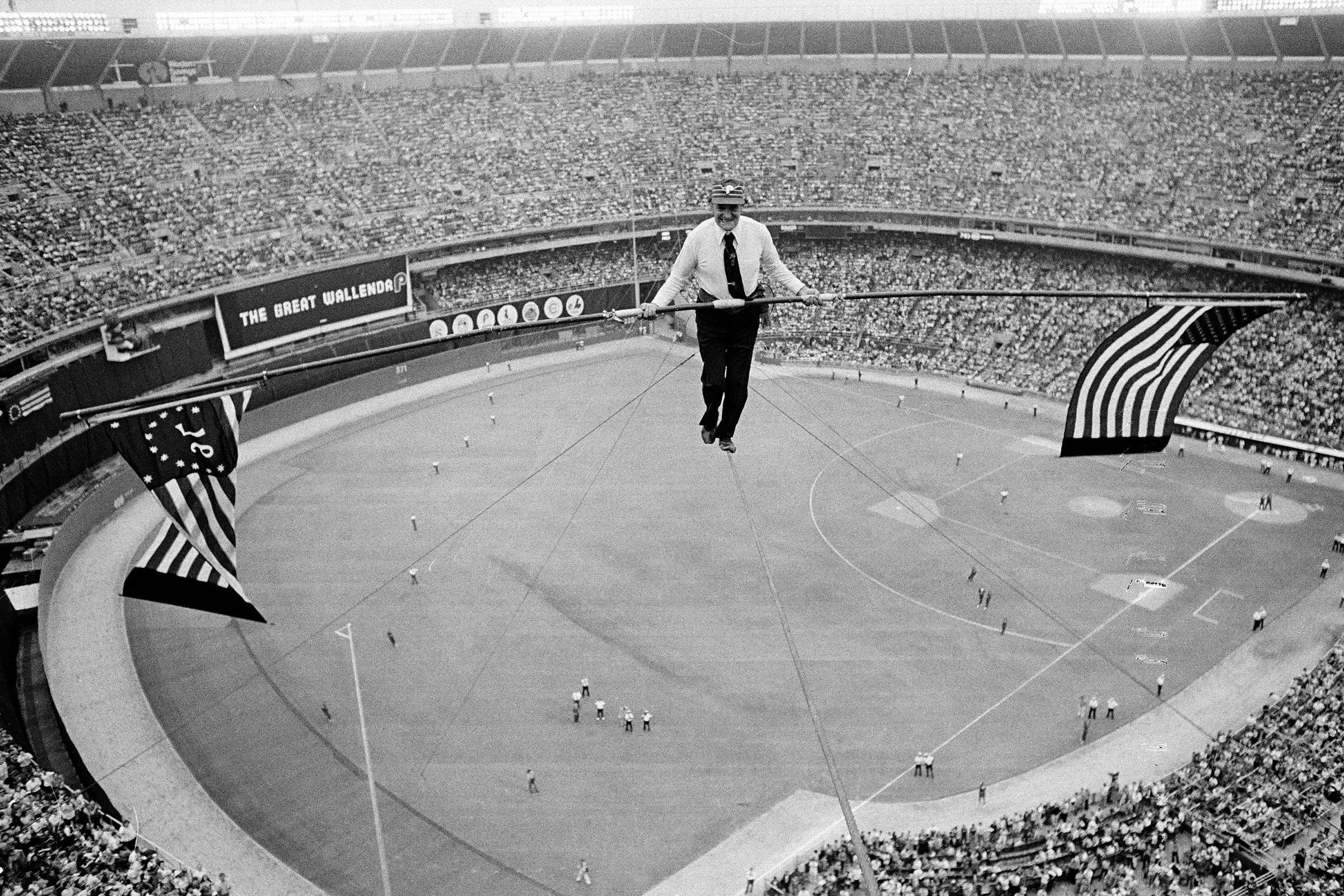 رجل يسير على حبل مشدود فوق ملعب مزدحم، يحمل أعلامًا أمريكية، في مشهد يعكس روح الوطنية والاحتفال.