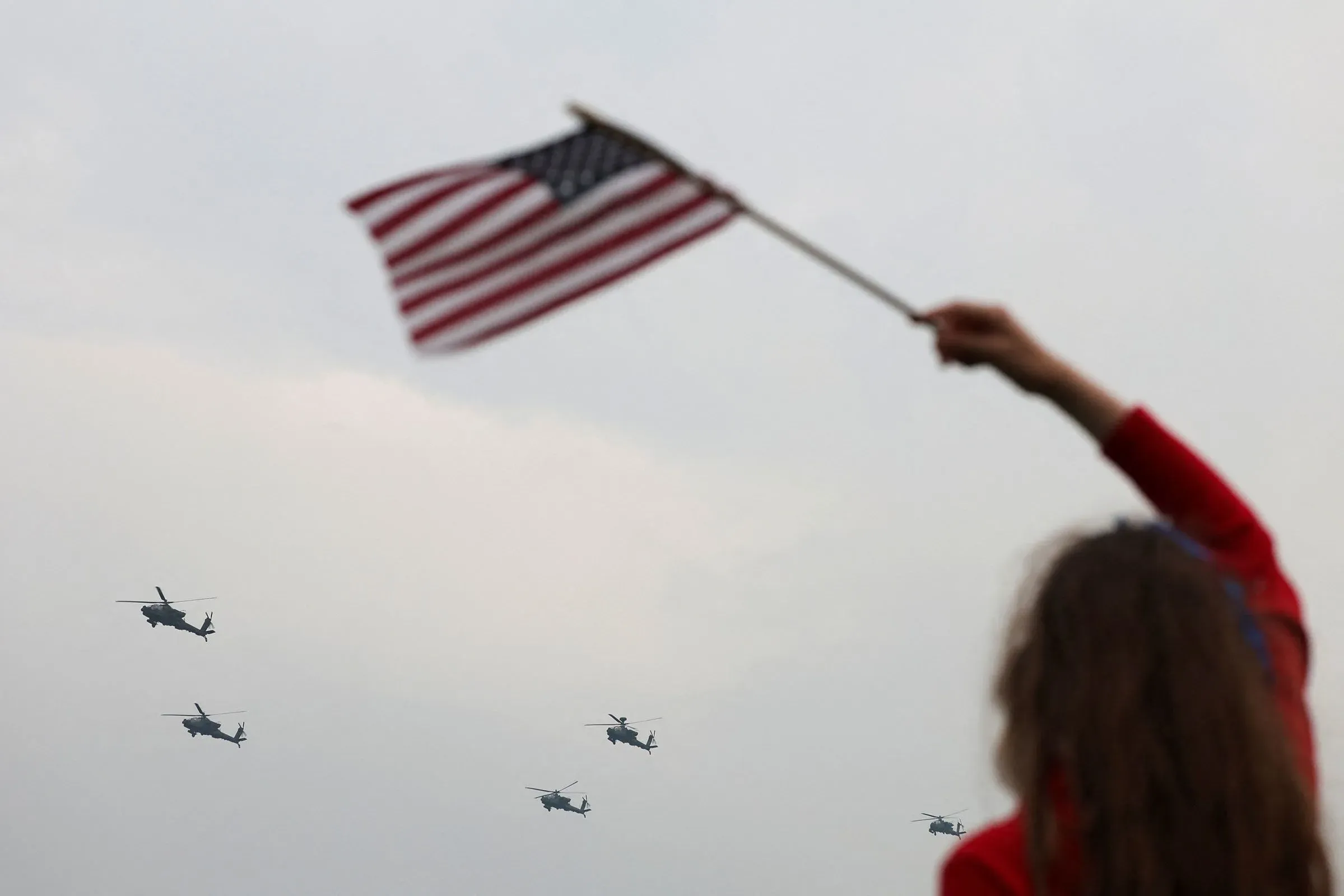 امرأة تحمل علم الولايات المتحدة بينما تحلق طائرات هليكوبتر في السماء، مما يعكس أجواء الاحتفالات الوطنية.