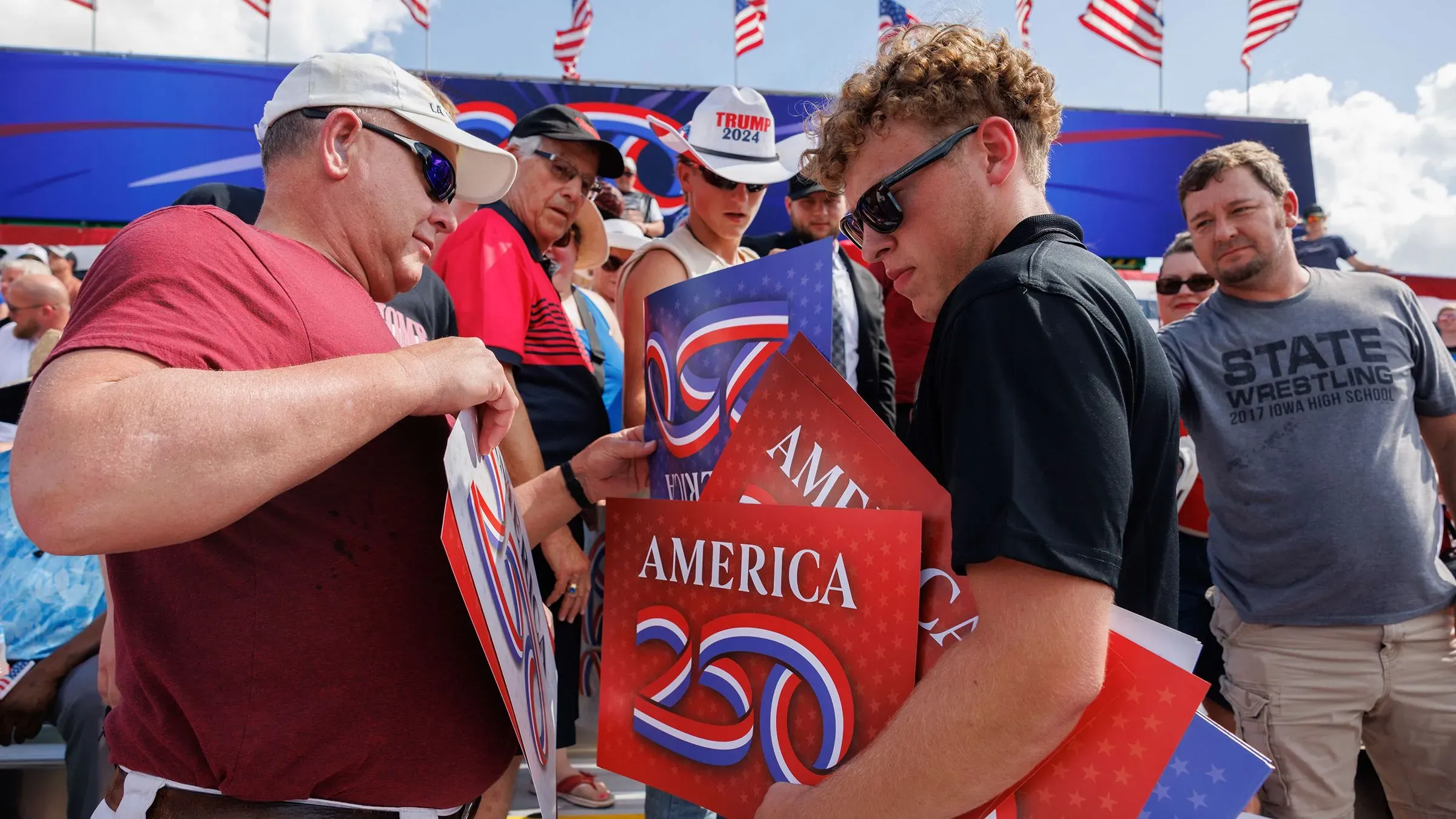 مجموعة من الأشخاص يحملون لافتات تحمل شعار "أمريكا 250" خلال حدث انتخابي لدونالد ترامب، مع أعلام أمريكية ترفرف في الخلفية.