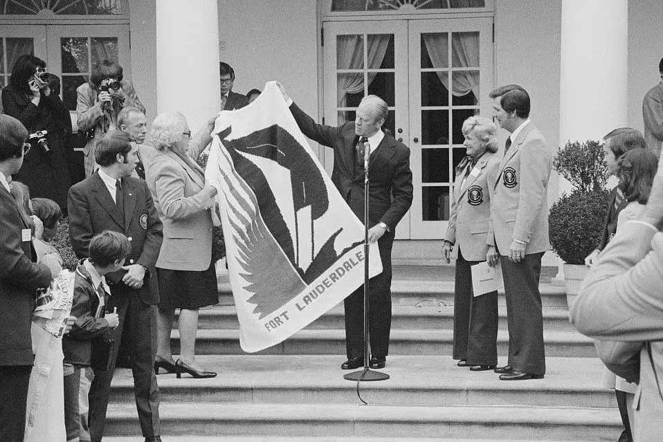 احتفال في البيت الأبيض حيث يظهر الرئيس جيرالد فورد وهو يعرض علم فورت لودرديل، مع حضور عدد من الشخصيات المحلية والإعلام.