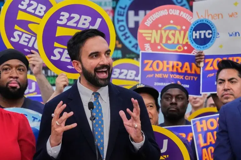 زهران ممداني يتحدث خلال حملته الانتخابية في نيويورك، محاطًا بأنصاره الذين يحملون لافتات تدعمه وتروج لبرامجه الاقتصادية.