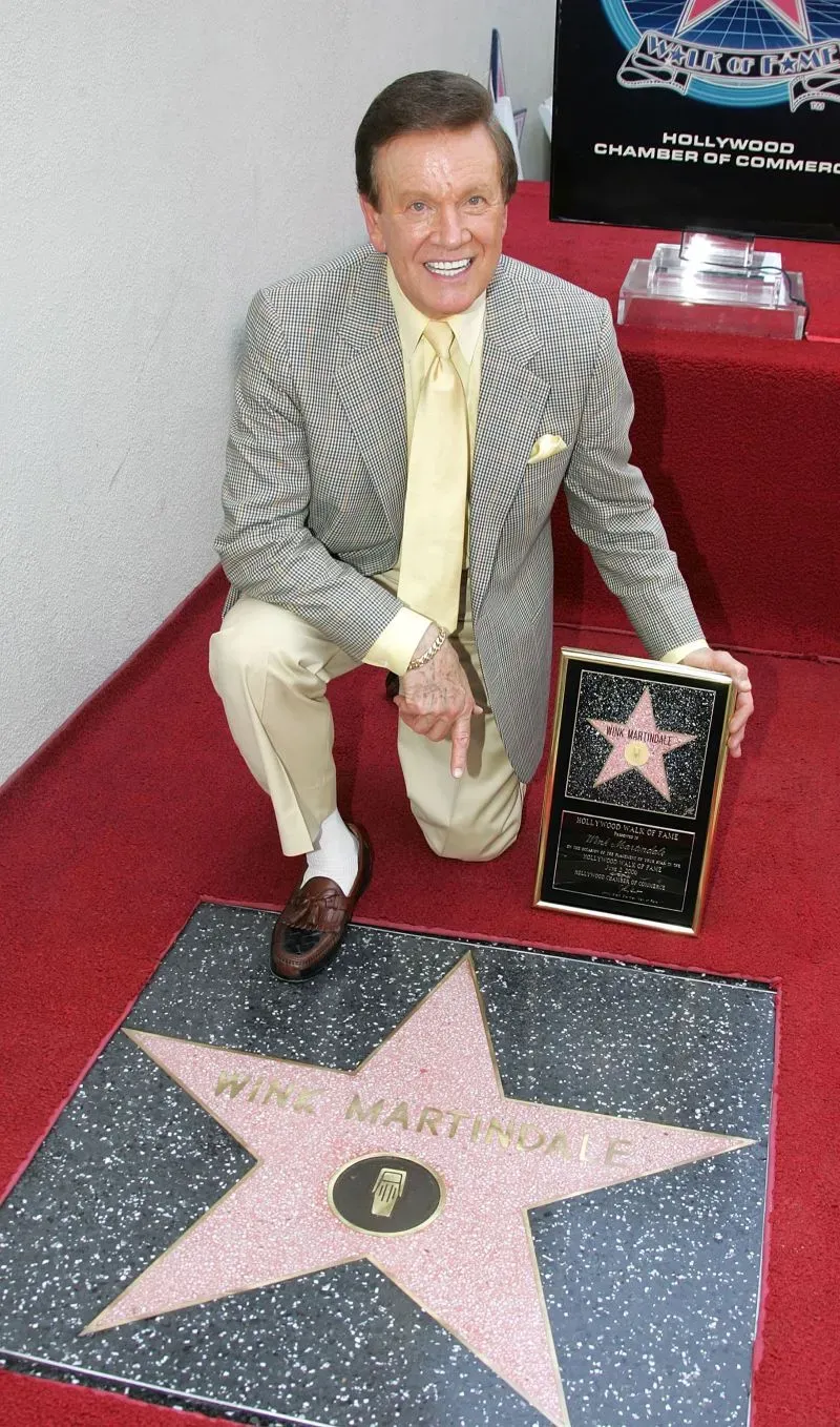 وينك مارتينديل يجلس بجانب نجمة على ممشى المشاهير في هوليوود، ممسكًا بشهادة تكريمية، مع ابتسامة تعكس مسيرته الفنية الطويلة.