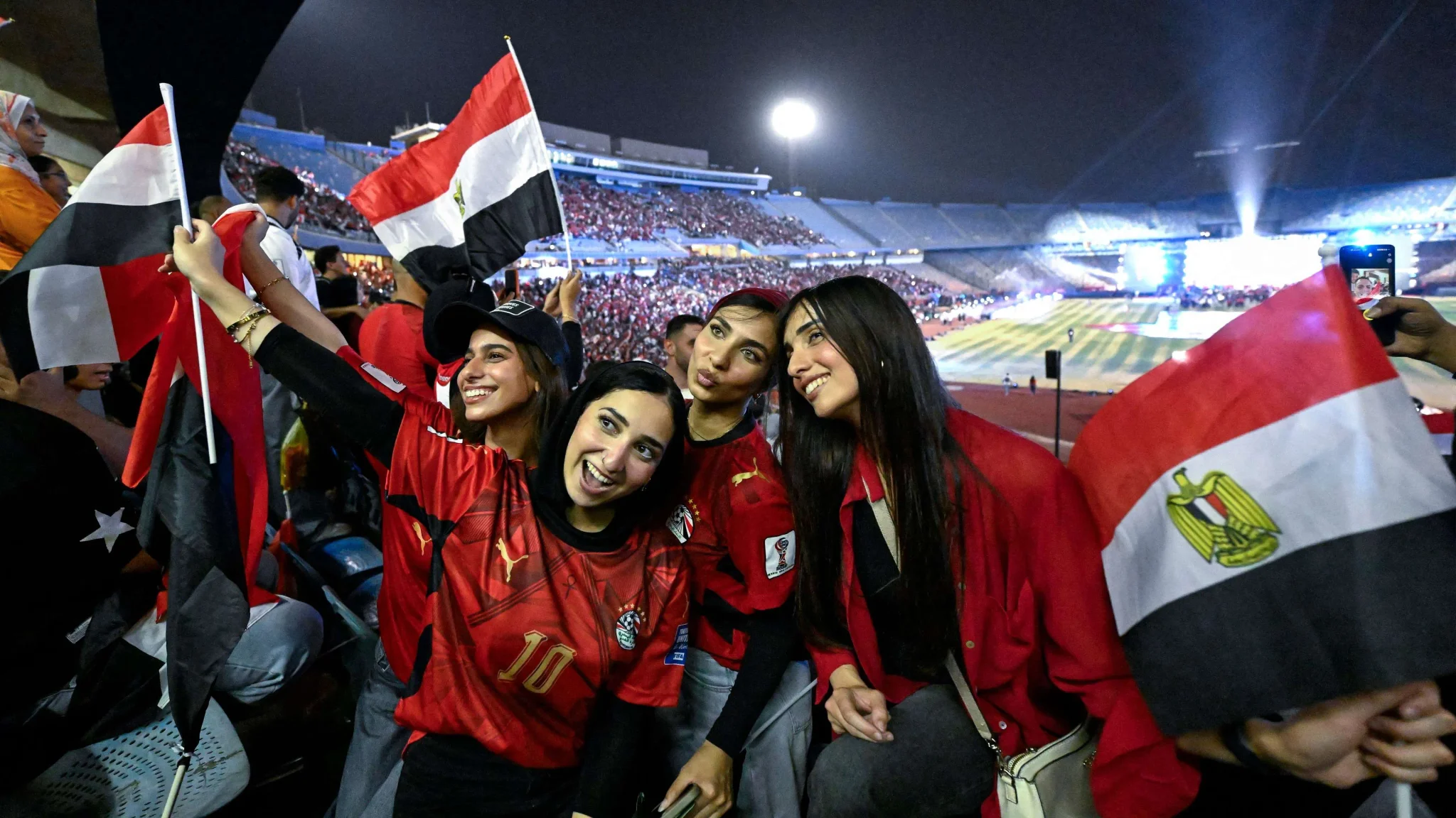 شابات مصريات يرفعن أعلام مصر ويحتفلن بحماس في استاد رياضي، معبّرات عن دعم منتخب مصر للسيدات تحت 18 سنة لكرة اليد.