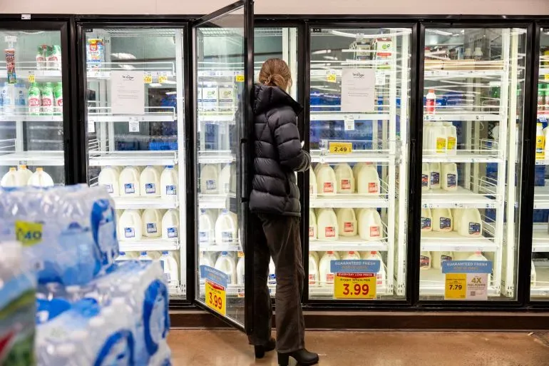 امرأة ترتدي سترة داكنة تقف أمام ثلاجات سوبرماركت، حيث تظهر أرفف الحليب شبه فارغة، مما يعكس الاستعدادات للعاصفة الشتوية.