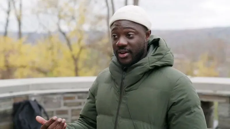 رجل يتحدث في مكان خارجي، يرتدي سترة خضراء وقبعة بيضاء، مع خلفية طبيعية تحتوي على أشجار صفراء، يعبر عن قلقه بشأن قضايا الهجرة.