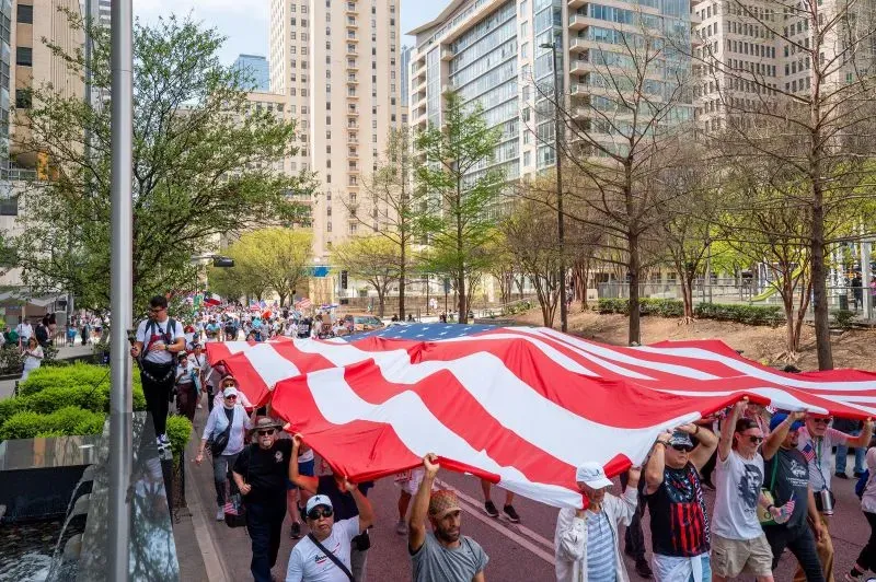 محتجون يحملون علمًا أمريكيًا كبيرًا أثناء مسيرة في دالاس، تعبيرًا عن المطالبة بإصلاح الهجرة وسط مخاوف من قمع المعارضة.