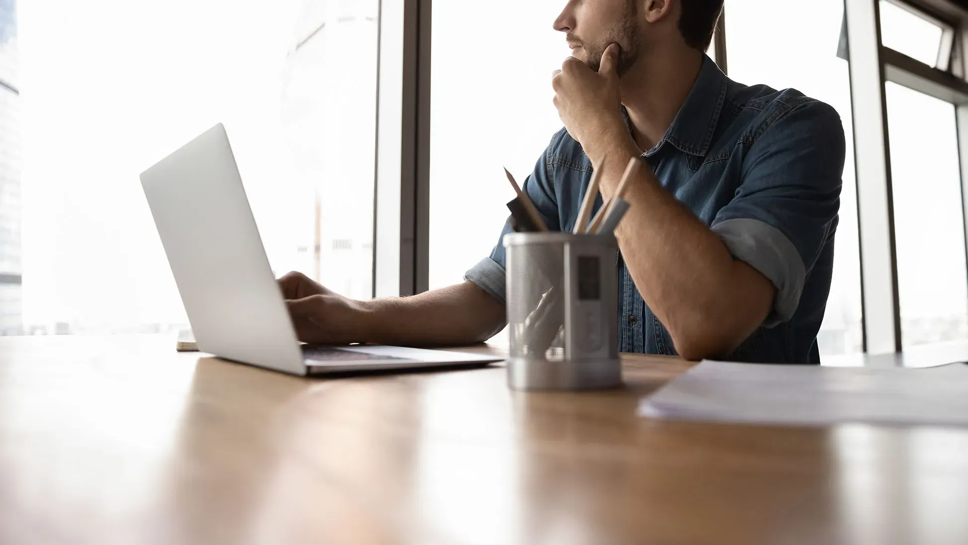 رجل يفكر أثناء استخدامه لجهاز كمبيوتر محمول على مكتب، مع أدوات مكتبية، في سياق الحديث عن تأثير الذكاء الاصطناعي على العمل.