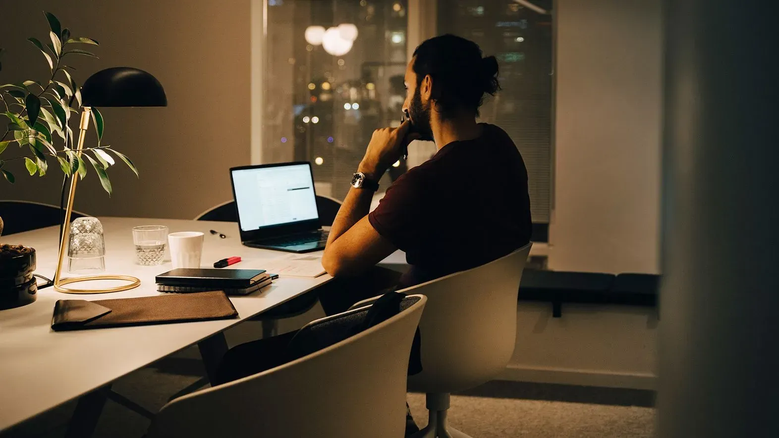 رجل يجلس بمفرده أمام كمبيوتر محمول في مكتب مضاء بشكل خافت، محاط بأدوات العمل، مما يعكس تأثير ساعات العمل الطويلة على الصحة العقلية.