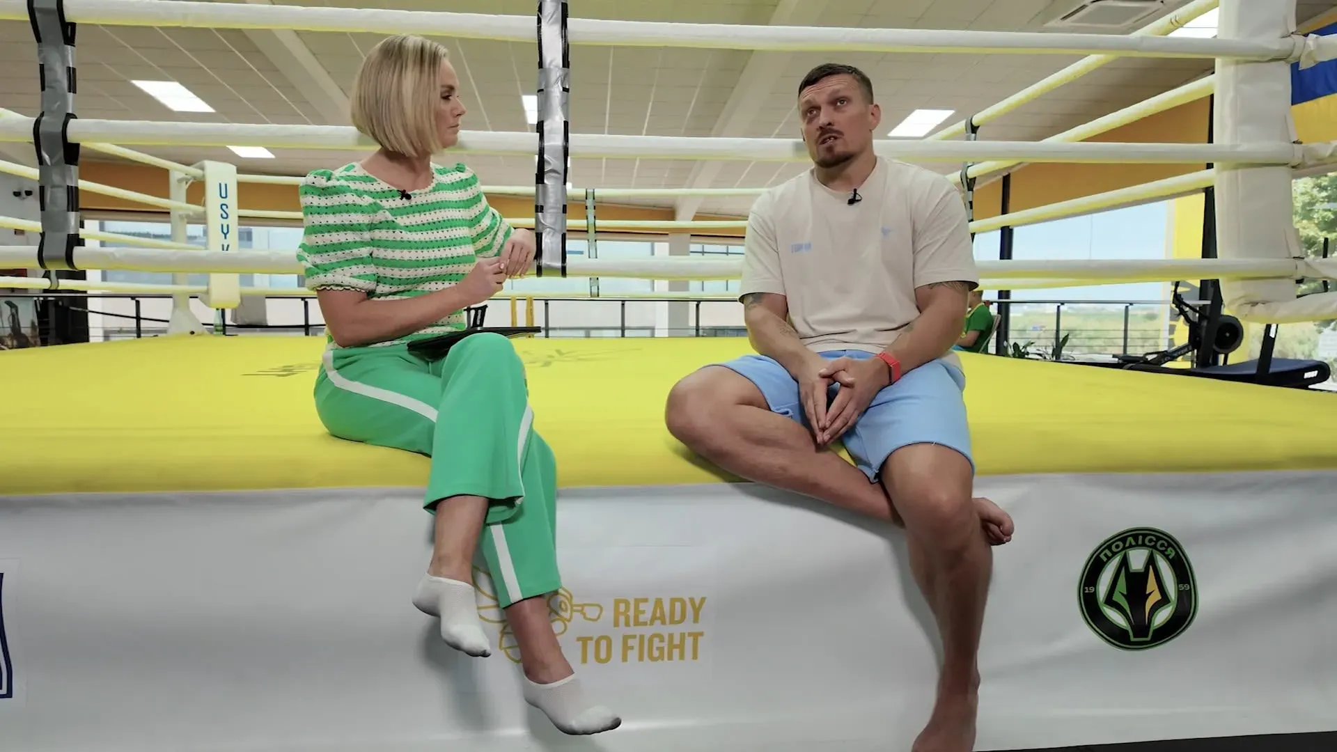 أولكسندر أوسيك، بطل العالم في الوزن الثقيل، يتحدث مع مراسلة في صالة رياضية، محاطًا بألوان العلم الأوكراني.