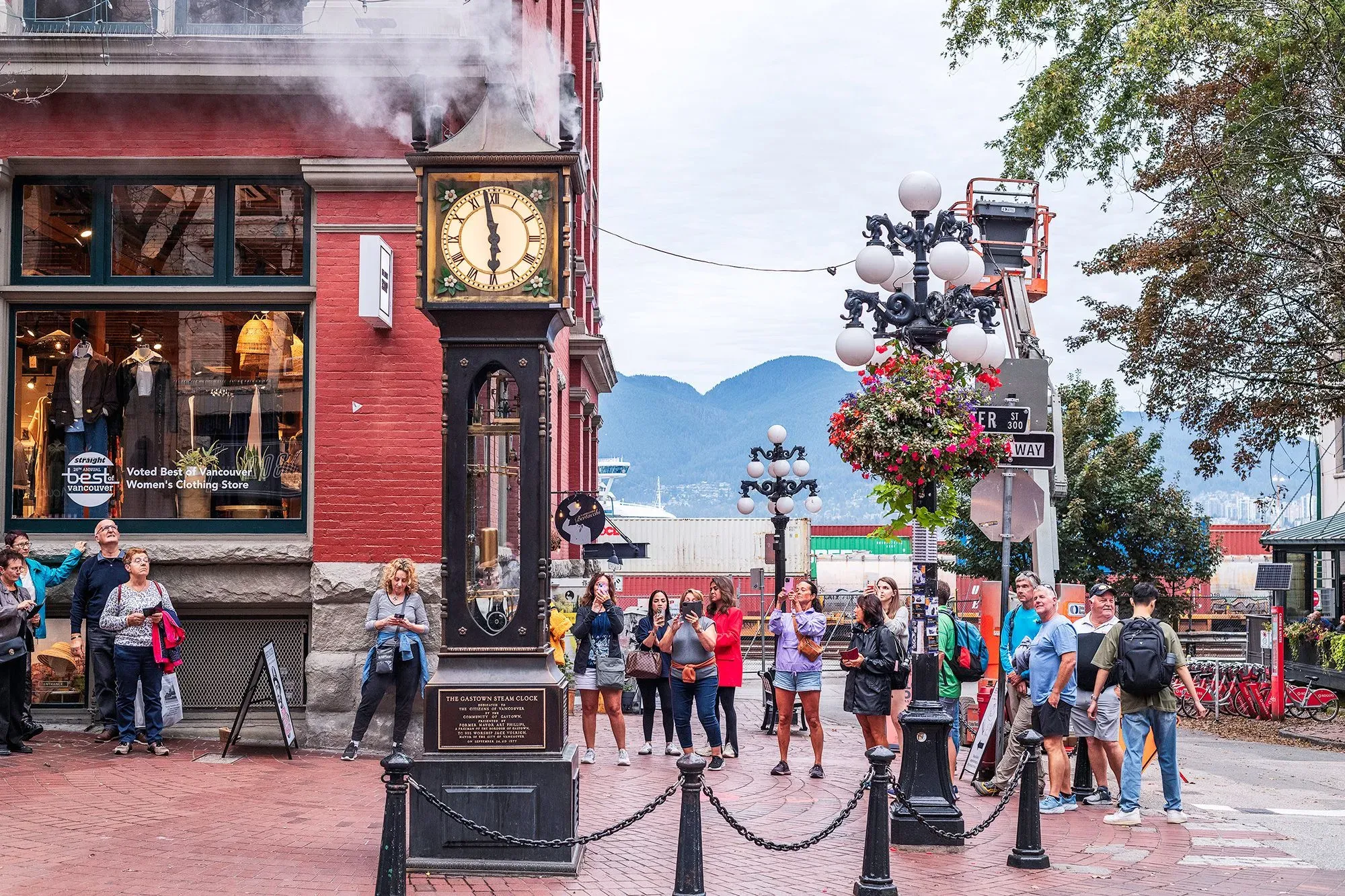ساحة مزدحمة في فانكوفر، حيث يظهر ساعة البخار الشهيرة والناس يتجمعون حولها، مع خلفية للجبال.