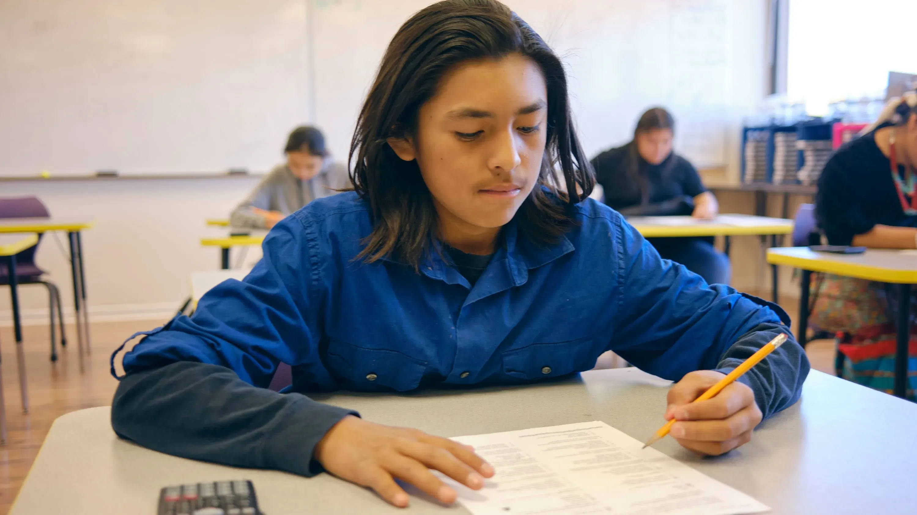 طالب يجلس في فصل دراسي، يعمل على اختبار مكتوب، مع التركيز على ورقة الاختبار. يبرز أهمية الأداء الأكاديمي للحصول على المساعدات المالية.