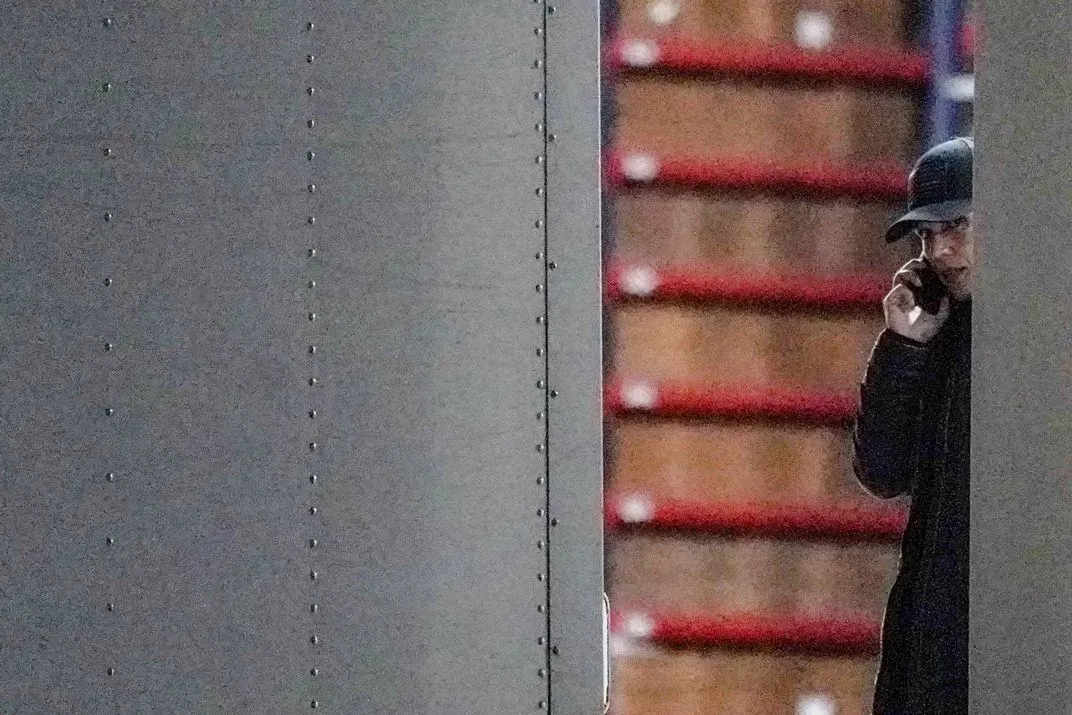 شخص يرتدي قبعة ويتحدث عبر الهاتف، يظهر جزئيًا خلف حائط معدني، مع خلفية تحتوي على صناديق خشبية، مما يعكس أجواء مرتبطة بالانتخابات.