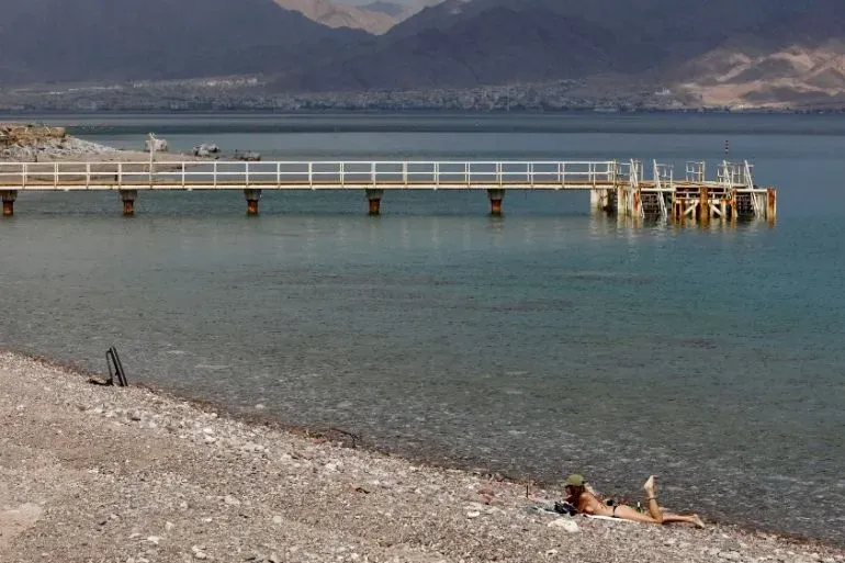 شاطئ مدينة إيلات يظهر رصيفًا خشبيًا يمتد نحو البحر الأحمر، مع شخص مستلقٍ على الرمال. الهجوم بالطائرات المسيرة من الحوثيين يثير القلق.