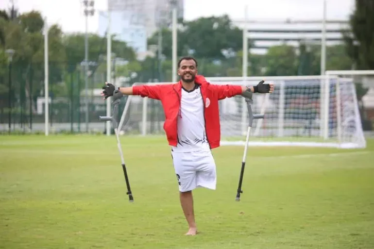 رجل مبتور الساقين يرتدي زي رياضي، يقف مبتسمًا على ملعب كرة القدم مع عكازين، مع خلفية مرمى، مما يعكس إرادة الحياة بعد تجربة صعبة.