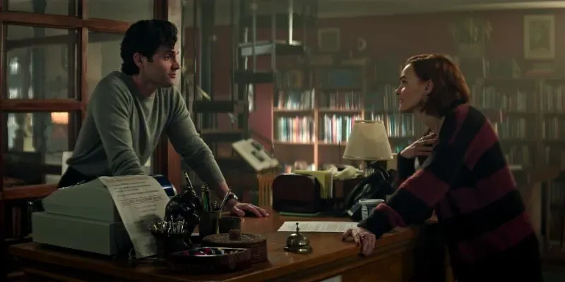 جو غولدبيرغ وكيت في مشهد من مسلسل "أنت"، يتحدثان في مكتبة، مع تفاصيل عن علاقة معقدة في سياق الحب والجريمة.