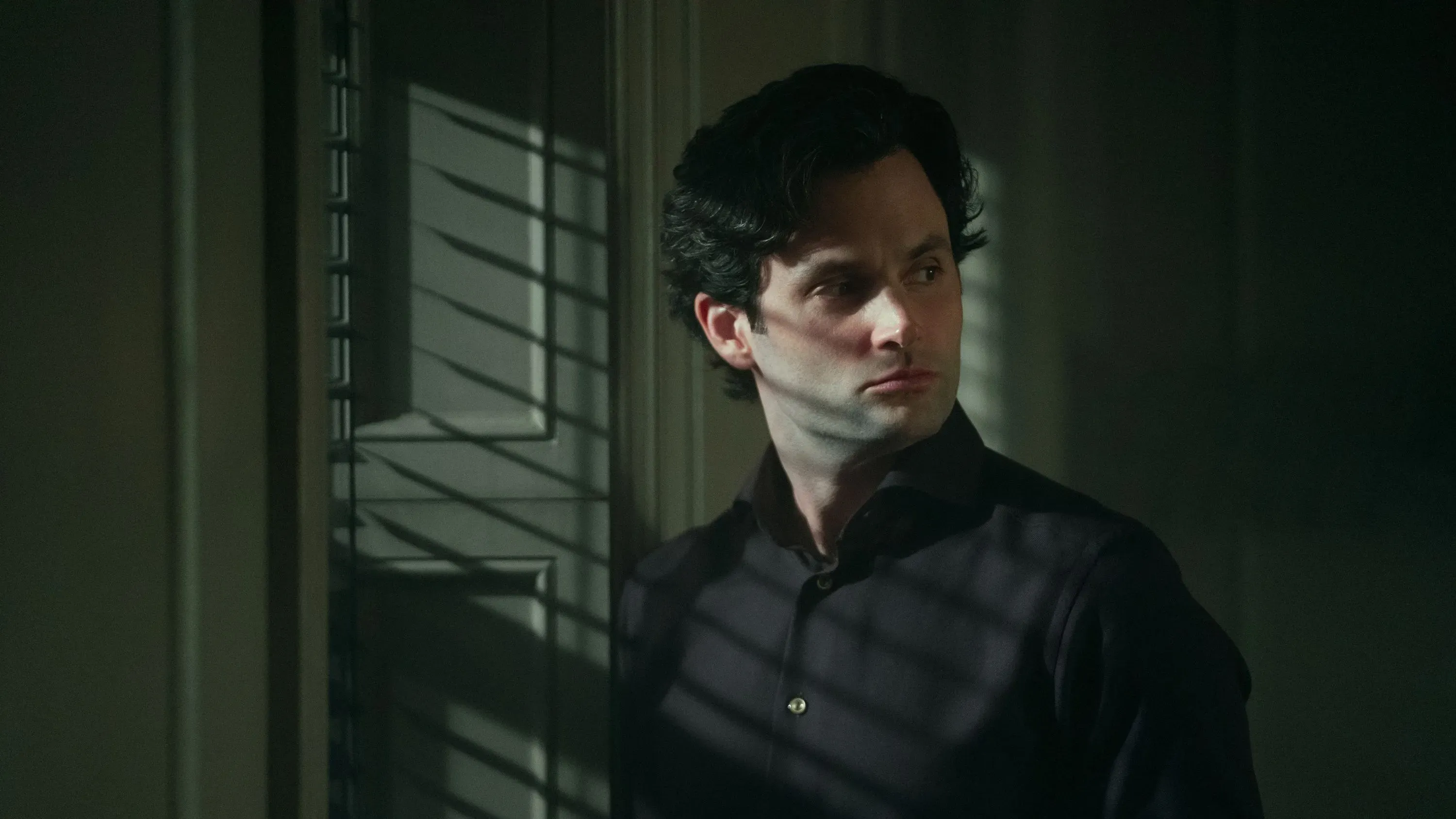 جو غولدبيرغ، الشخصية الرئيسية في مسلسل "أنت"، يقف في غرفة مضاءة بخطوط من الظل، مع تعبير وجه يعكس الصراع الداخلي.