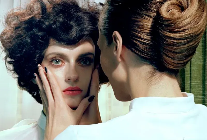 امرأة ذات شعر مجعد تتأمل في مرآة، بينما تلمس وجهها امرأة أخرى ذات تسريحة شعر أنيقة، في خلفية ذات ألوان دافئة.