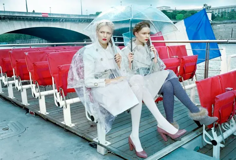 امرأتان ترتديان ملابس عصرية، واحدة تحمل مظلة شفافة، تجلسان على متن قارب مع مقاعد حمراء، خلفهما جسر ومياه.