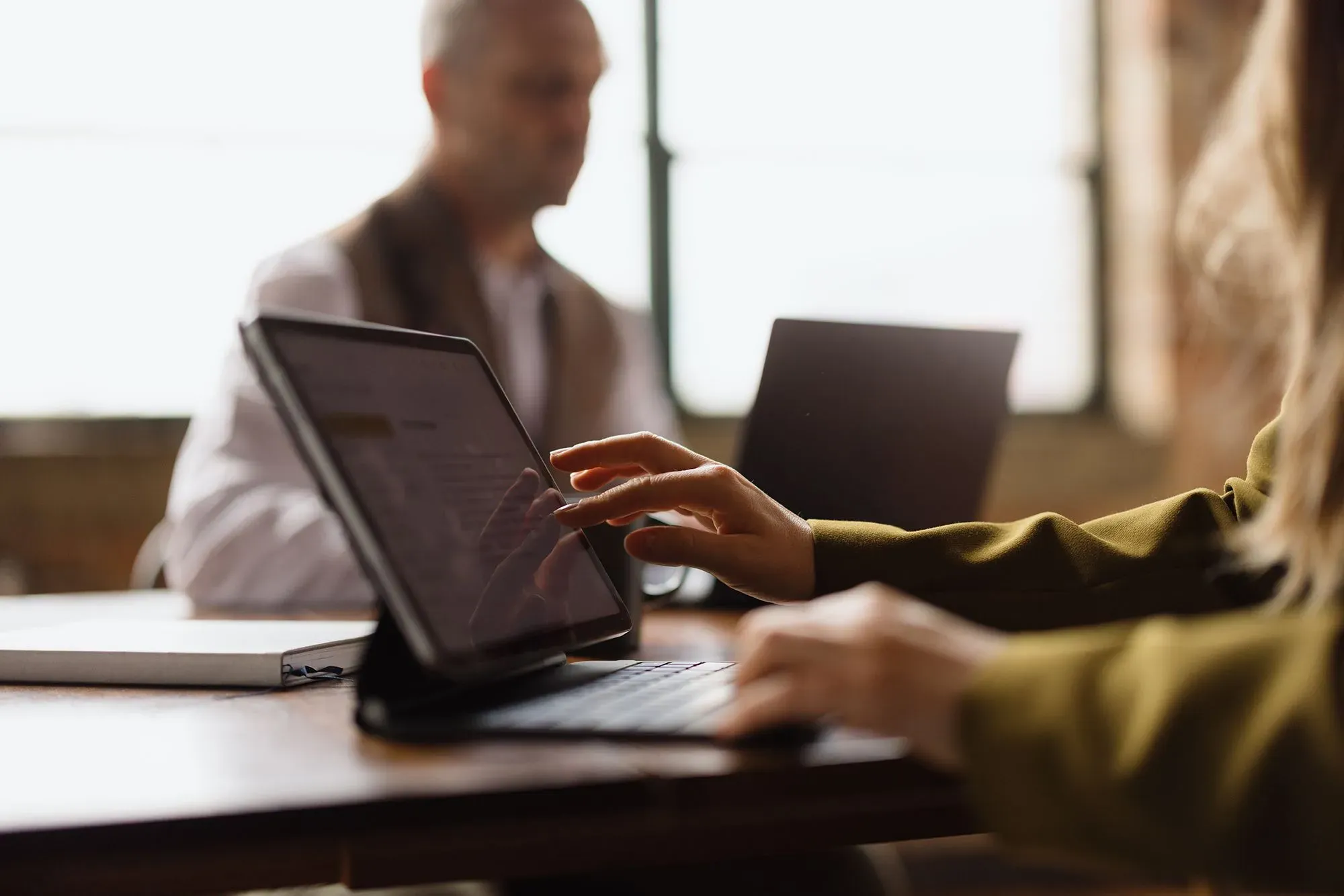 امرأة تستخدم جهاز لوحي للبحث أو العمل، بينما يظهر في الخلفية شخص آخر يعمل على جهاز كمبيوتر محمول، في سياق الاعتماد المتزايد على التكنولوجيا والذكاء الاصطناعي.