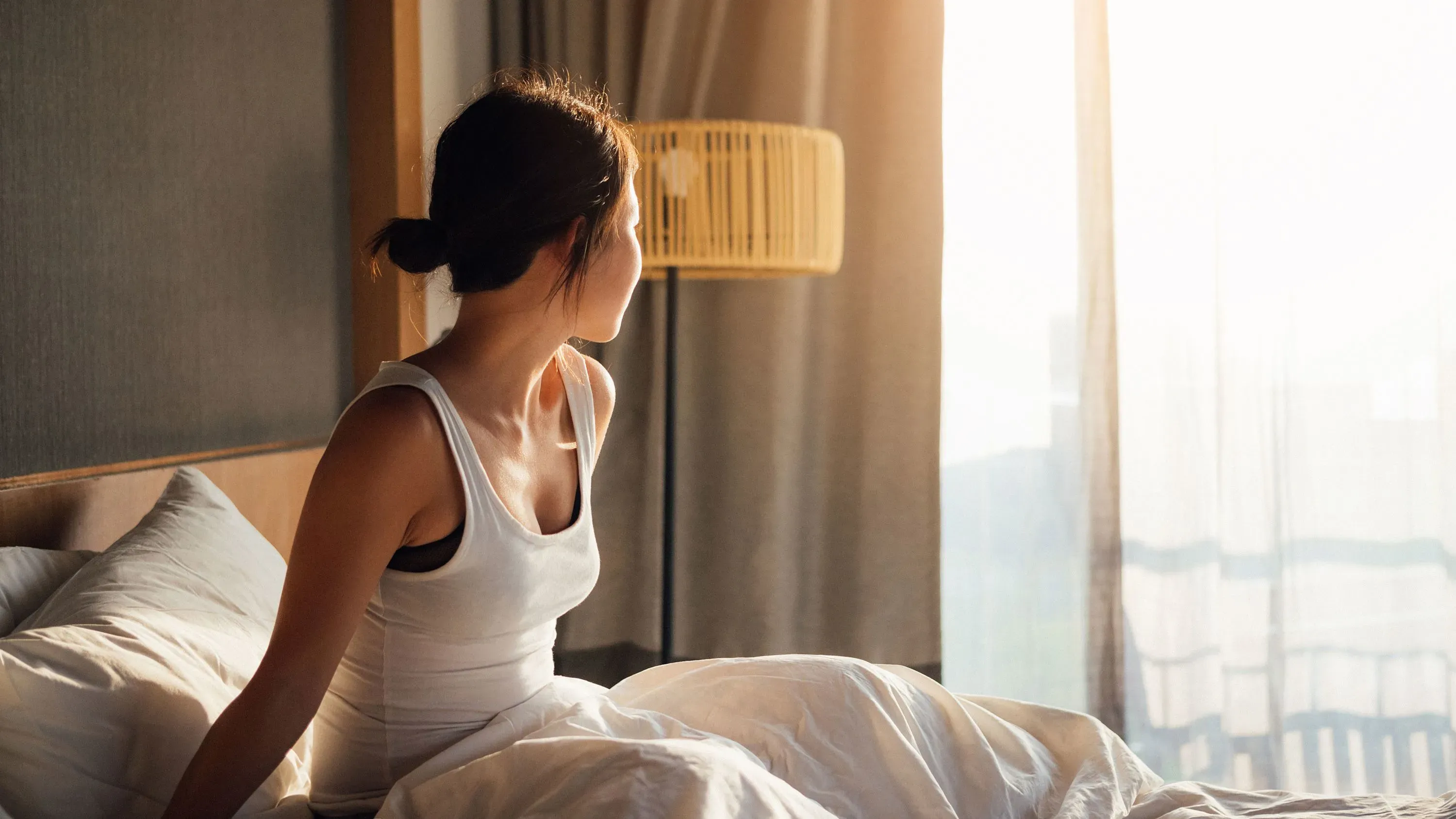 امرأة تجلس على السرير، تنظر نحو نافذة مضاءة بأشعة الشمس، تعكس أهمية النوم للصحة النفسية والرفاهية.