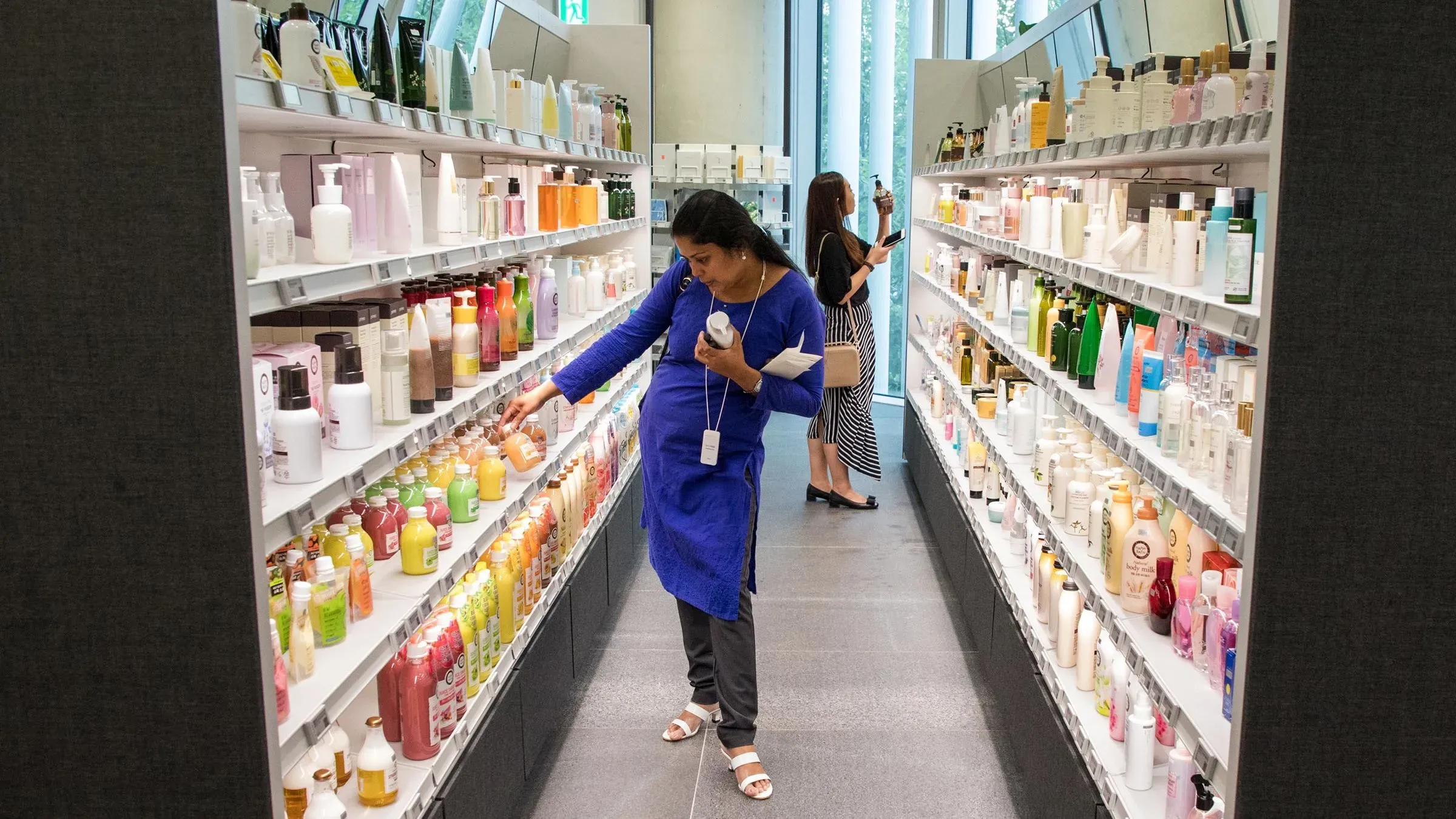 امرأة تتسوق في متجر لمنتجات التجميل الكورية، محاطة برفوف مليئة بزجاجات مستحضرات العناية بالبشرة، مع زبائن آخرين في الخلفية.