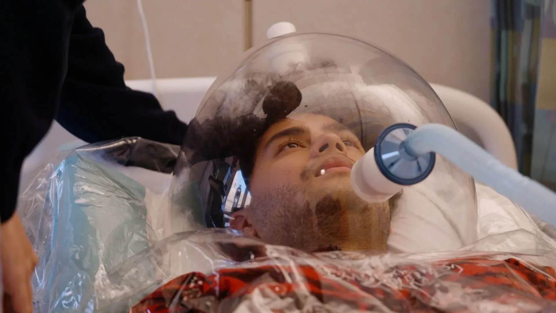 شاب يتلقى علاجًا طبيًا، محاط بكبسولة شفافة، مع أنبوب توصيل للهواء. تظهر تفاصيل تكنولوجية حديثة في بيئة طبية.