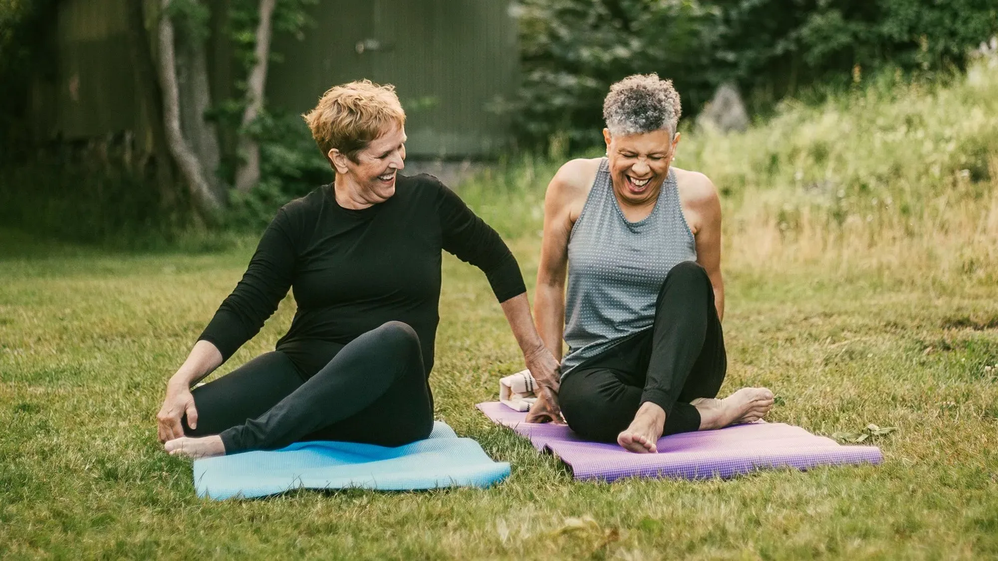 امرأتان يجلسان على حصيرتين في حديقة، يضحكان ويتبادلان الأحاديث أثناء ممارسة اليوغا، مما يعكس أهمية الروابط الاجتماعية للصحة النفسية.